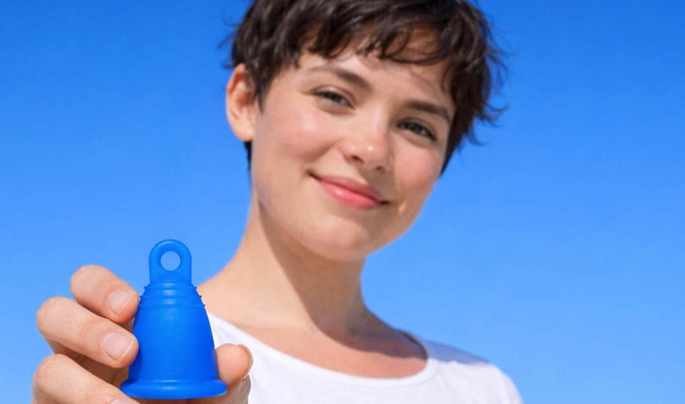 A woman with short dark hair holds a bright blue silicone menstrual cup against a clear blue sky background.